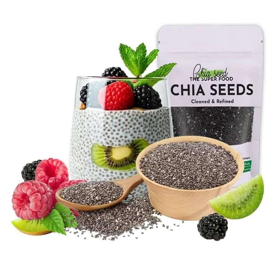 Glass of chia seed pudding topped with fresh berries and mint, surrounded by a packet of chia seeds, a bowl and spoonful of chia seeds, and assorted fruits like kiwi, raspberry, and blackberry.